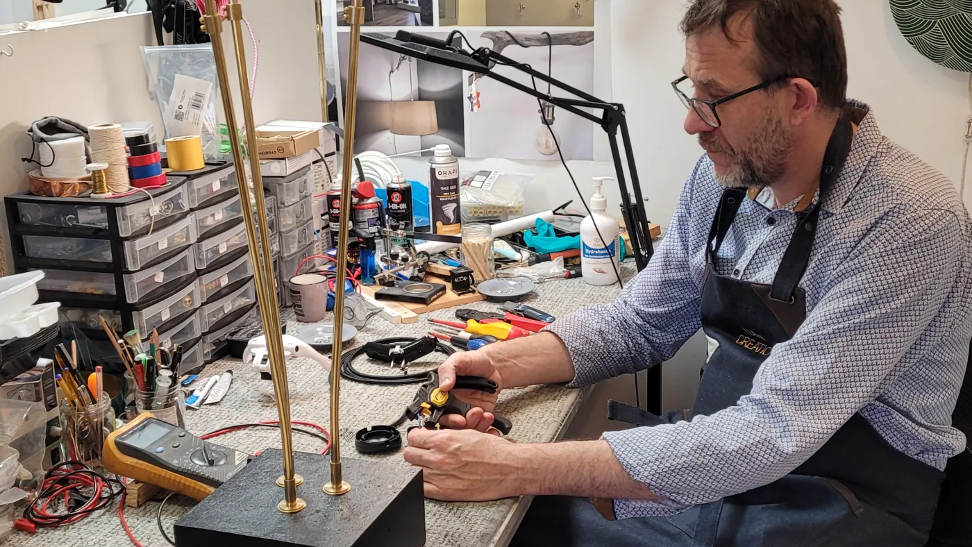 Dans l'atelier Lumix Création : montage d'un luminaire à tiges laiton sur socle noir, un contrôleur d'isolement posé sur l'établi.